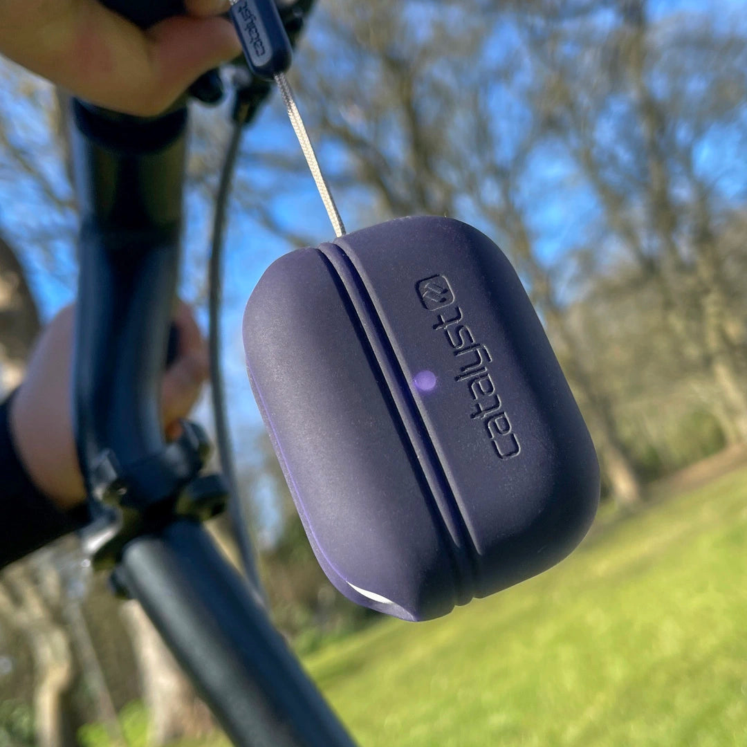 An Ink-colored AirPods Pro - Essential Case by Catalyst, crafted from silicone, hangs from a bicycle handlebar in a park setting. With trees and grass in the background, this drop proof accessory adds style while protecting from scratches.