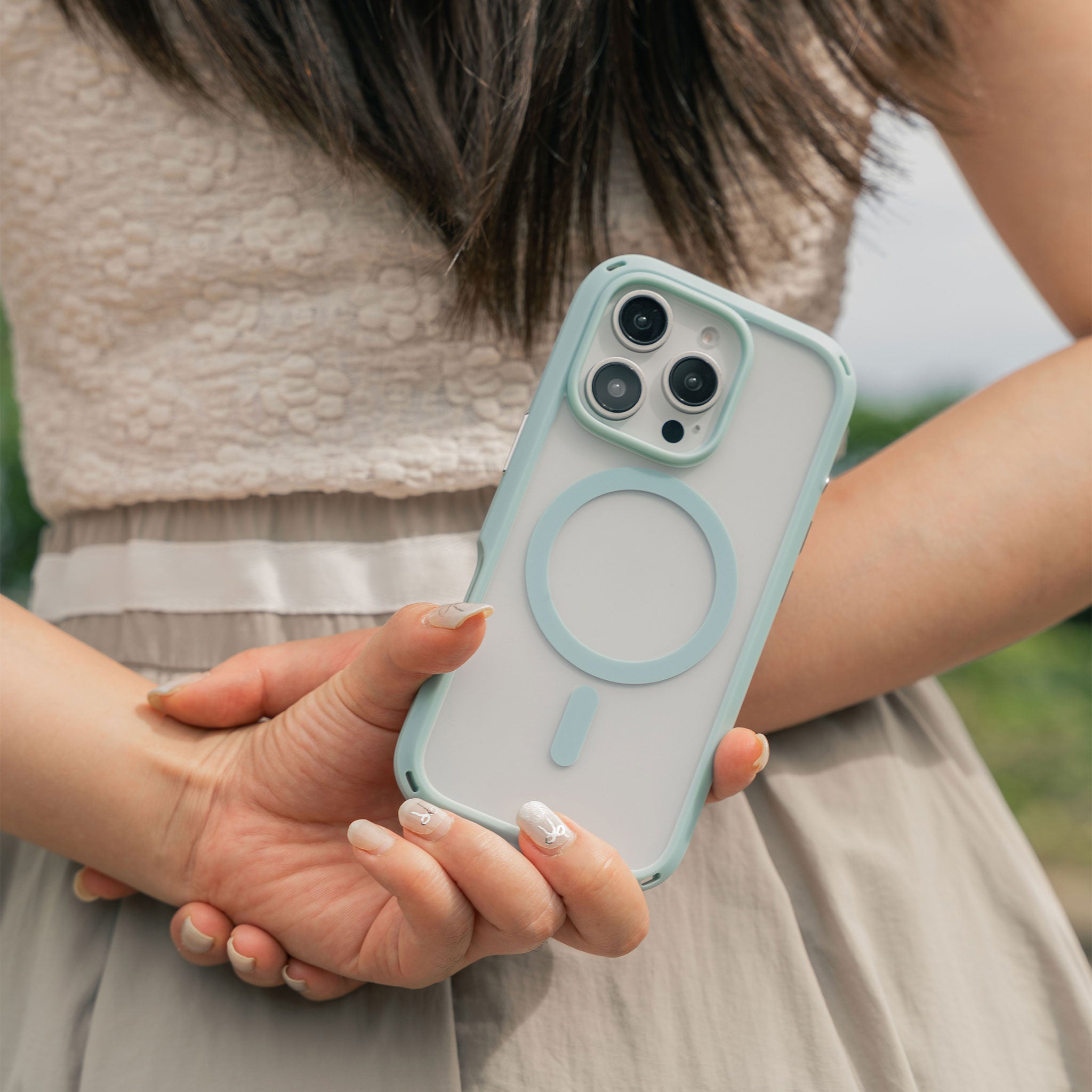 A person is holding a smartphone with a Catalyst Drop Proof Case in Sky Blue behind their back. The phone features a three-camera array. The background is blurred, and the person is dressed in a white top and light gray skirt.