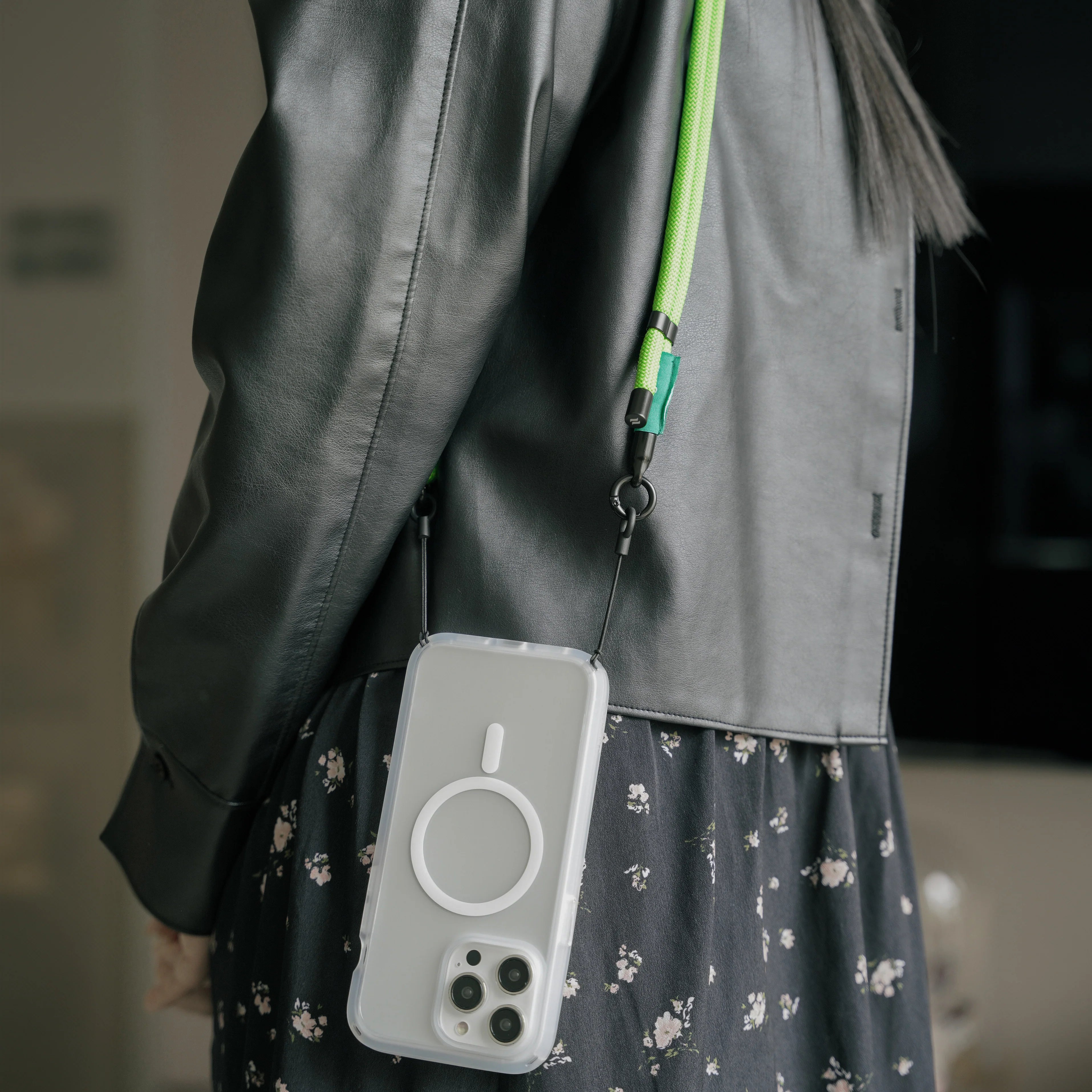 A person in a black jacket carries an iPhone on a green Catalyst Crossbody Shoulder Strap, featuring a circular design. The phone is securely attached to its clear case and worn crossbody for easy access.