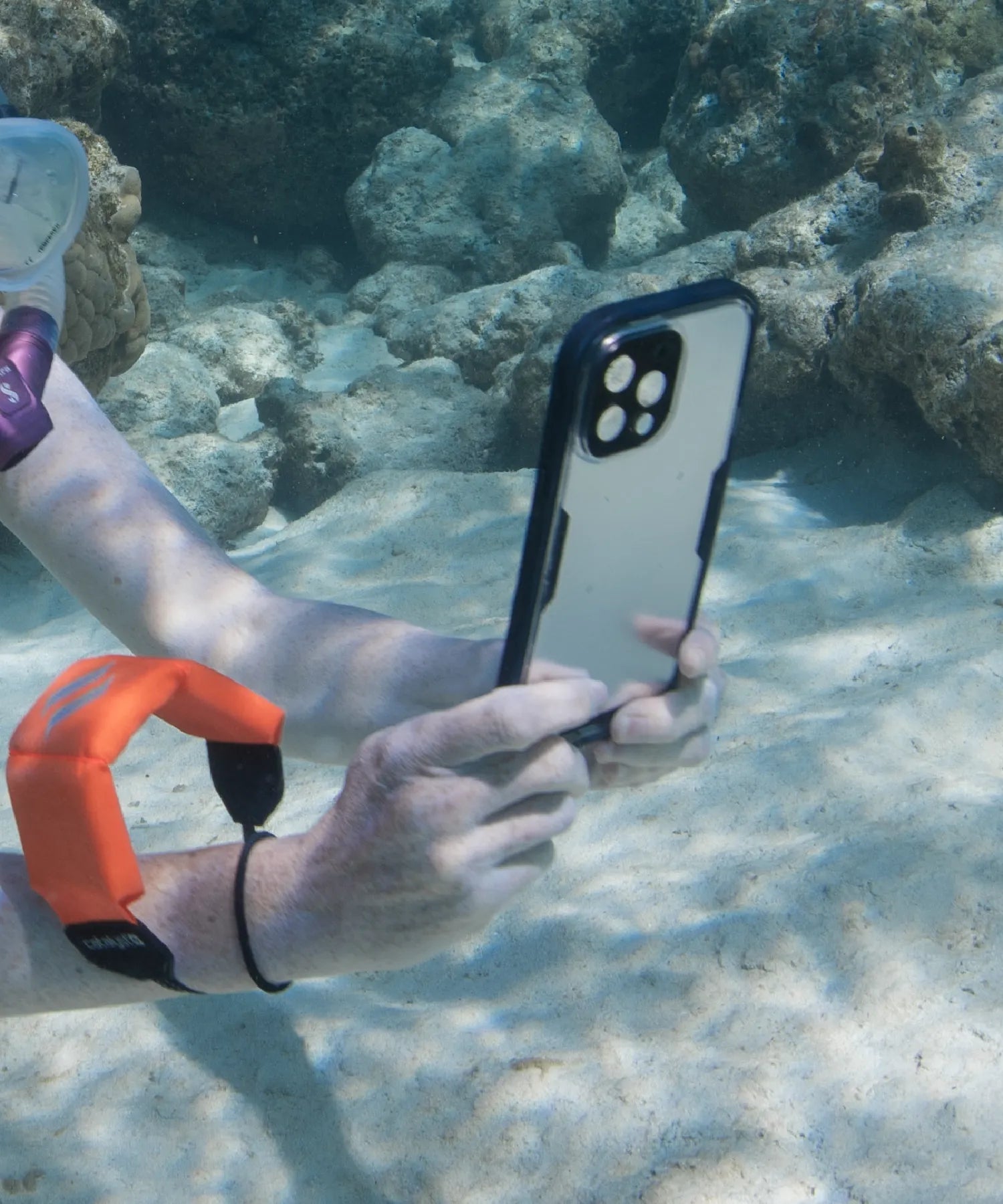 A close up of a woman taking a photo underwater in a black Catalyst waterproof case. The iPhone is secured to her wrist through a Floating Lanyard in orange. 