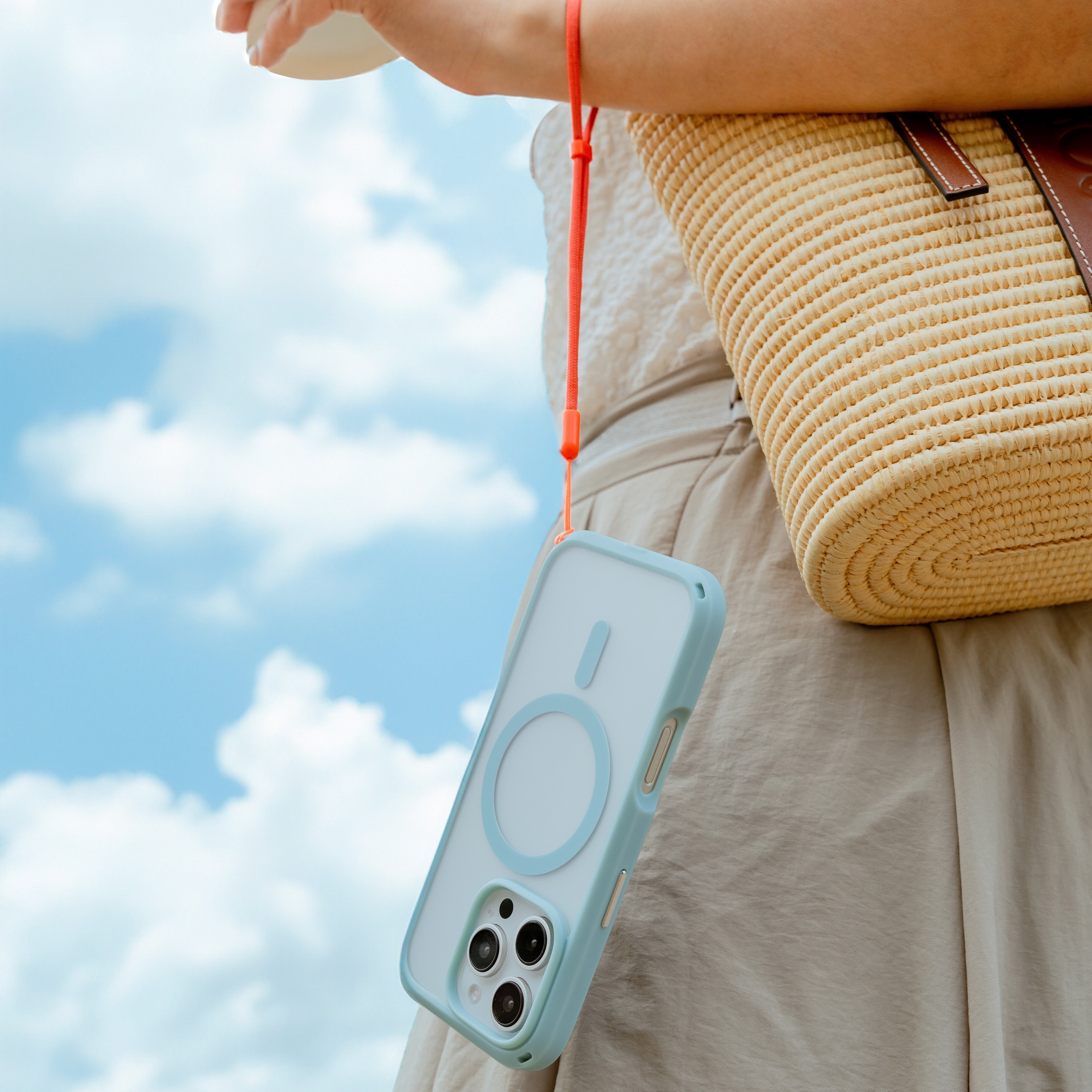 Woman holding a phone encased in the Sky Blue Drop Proof Case by Catalyst, attached to a wrist strap, carrying a woven bag against a backdrop of a cloudy sky.